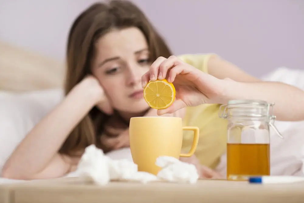 Woman, laying down, squeezing lemon into her mug next to a pile of tissues, honey and a thermometer